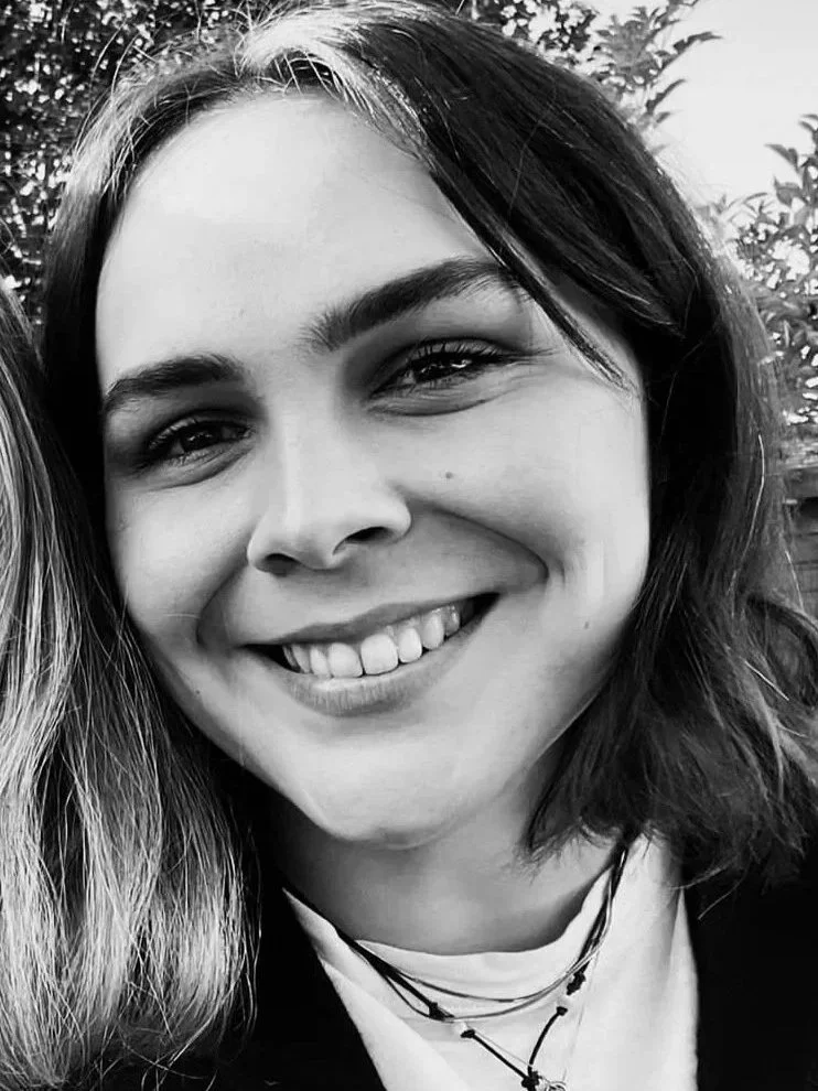 Close-up of a young woman smiling with visible teeth, dark hair, and expressive eyes, outdoor background with trees.