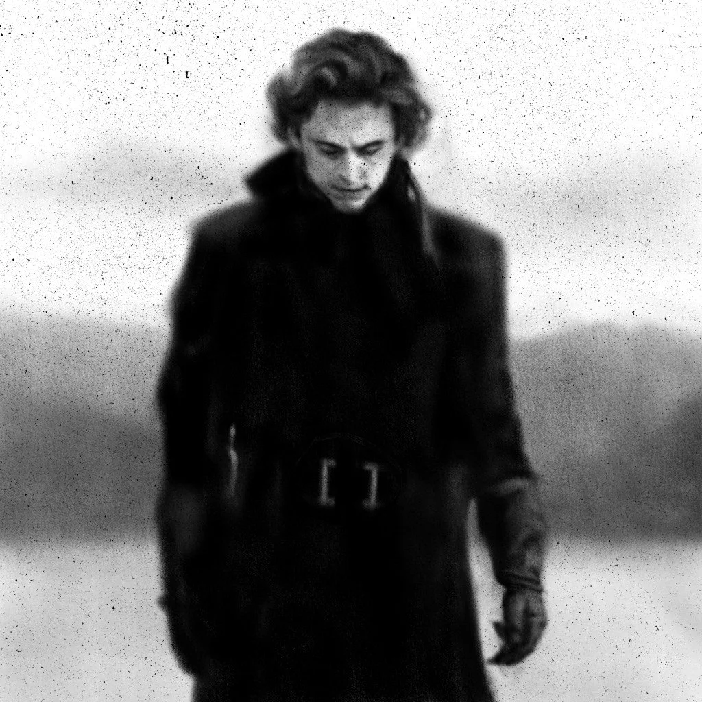 Black and white photo of a woman with short, curly hair, looking downward, wearing a dark coat with a high collar and gloves, outdoors with a blurry landscape in the background.