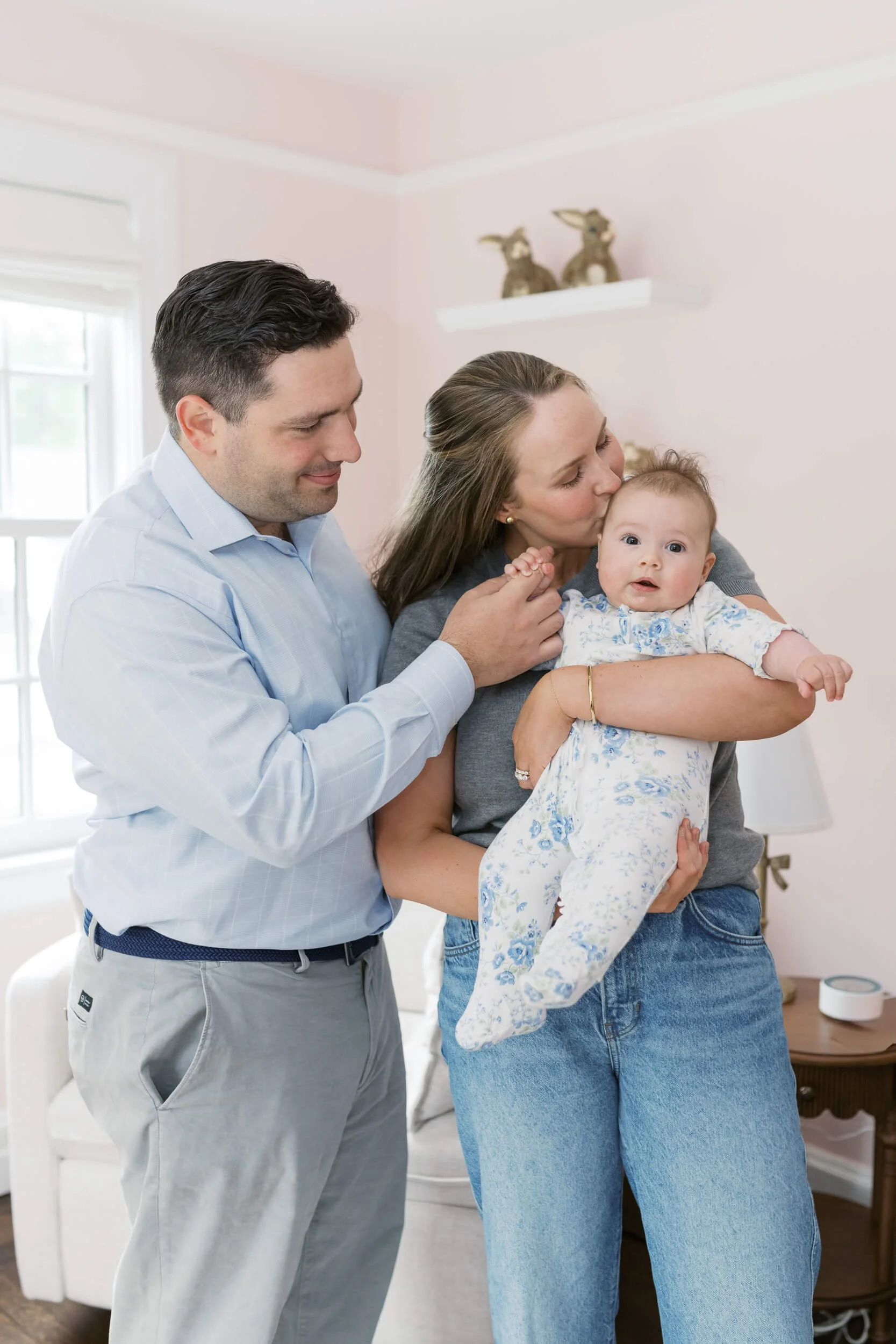 Family portrait in a soft pink nursery, parents holding their daughter during her milestone photos on Long Island.