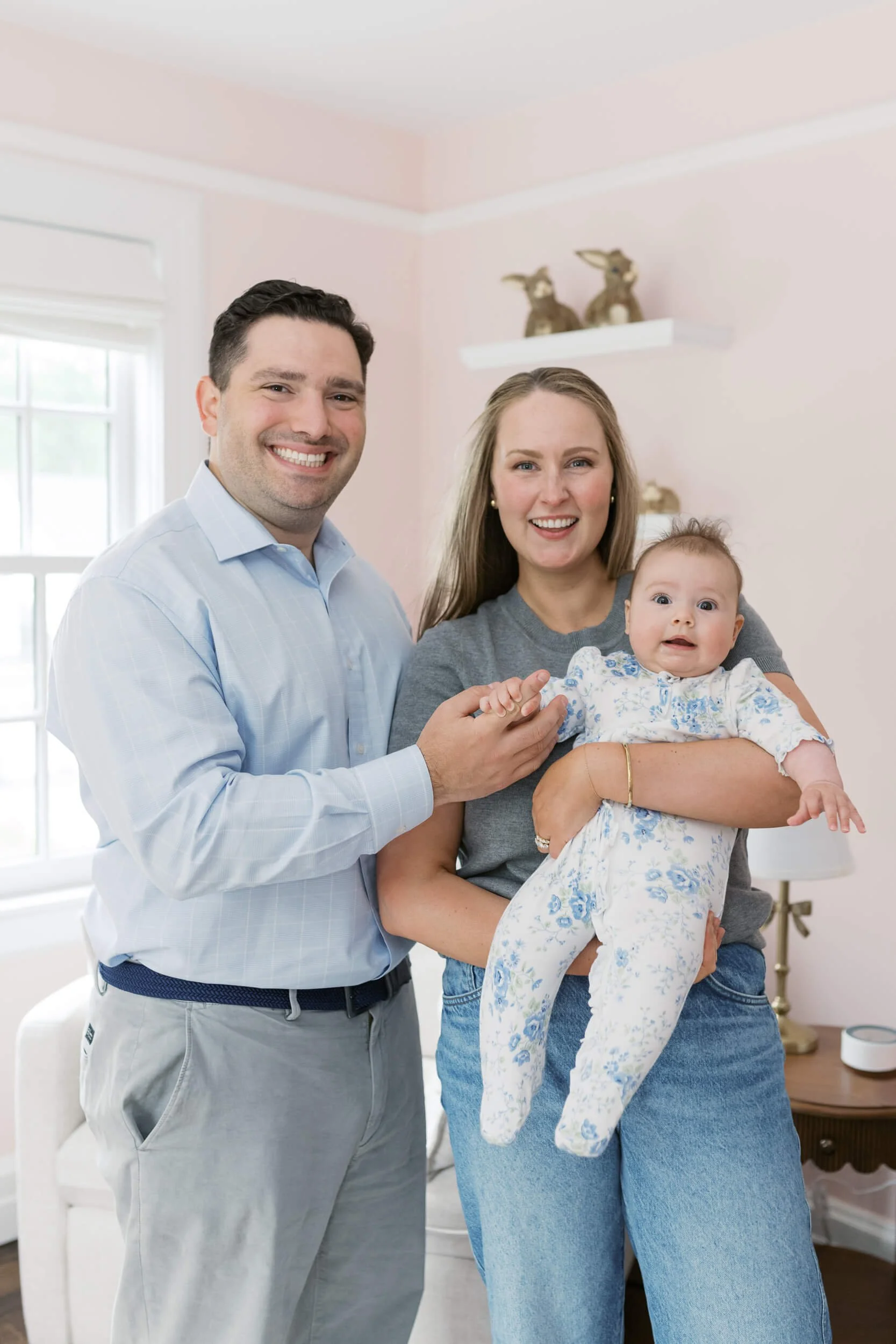 Family portrait in a soft pink nursery, parents holding their daughter during her milestone photos on Long Island.