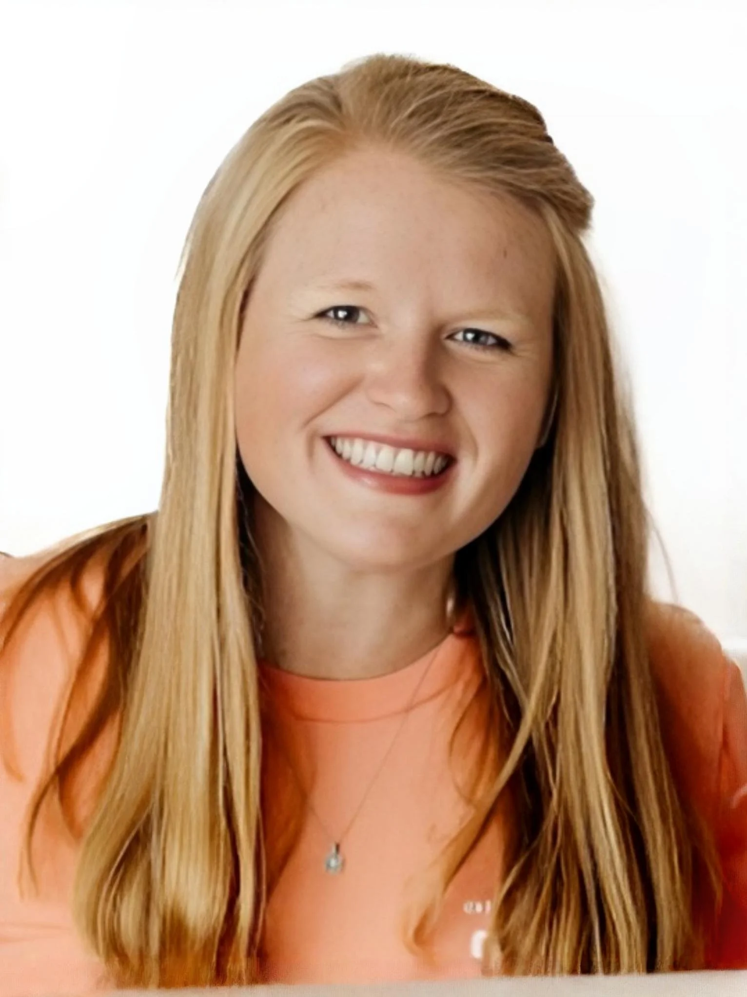 A young woman with long blonde hair, smiling, wearing an orange top, against a plain white background.