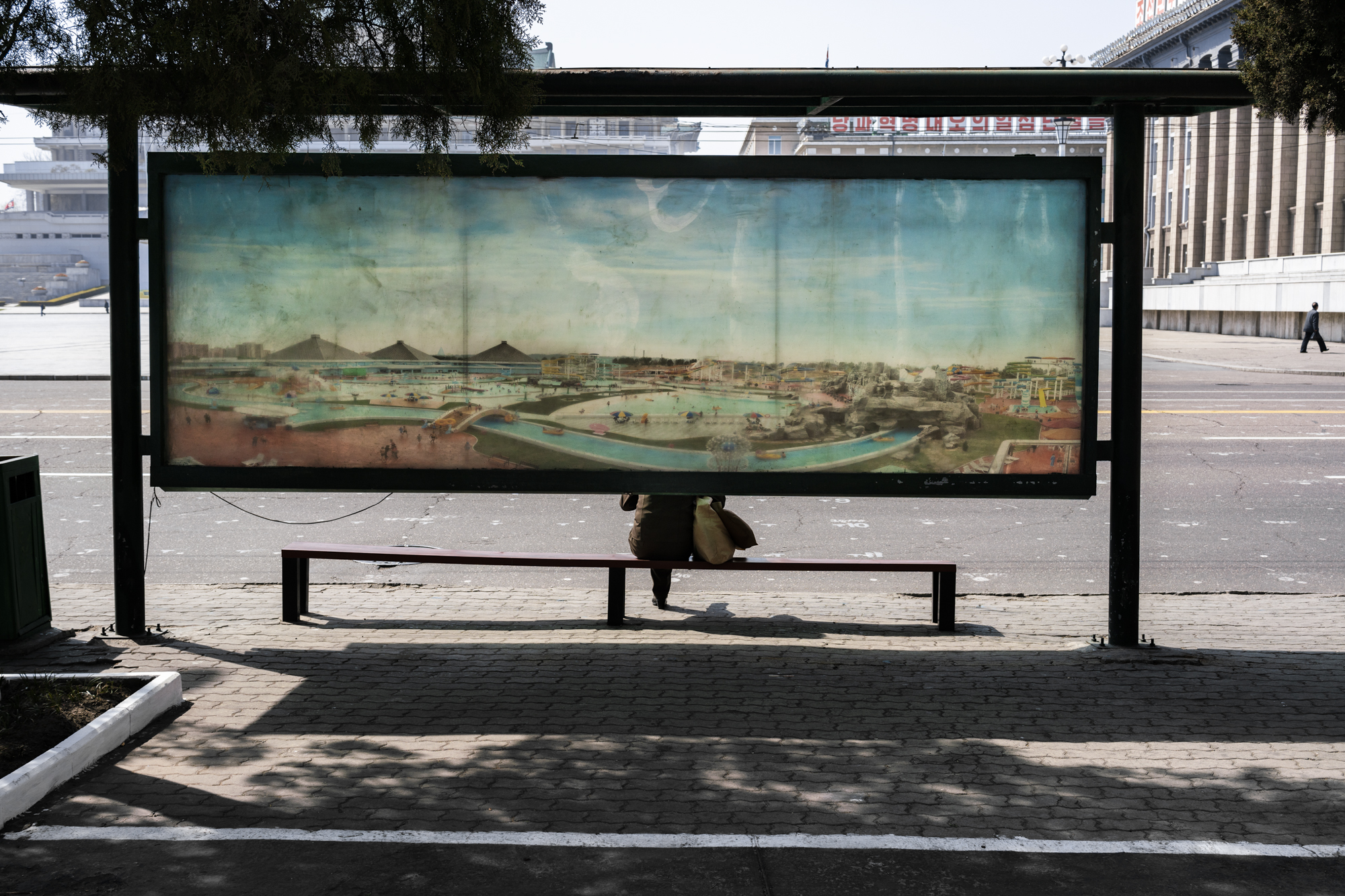 Street scene. A woman is waiting for a bus in Kim Il Sung Square. The back of the bus stop itself is a large-scale print of the Munsu Water Park, a state run water park located in the east of Pyongyang.
