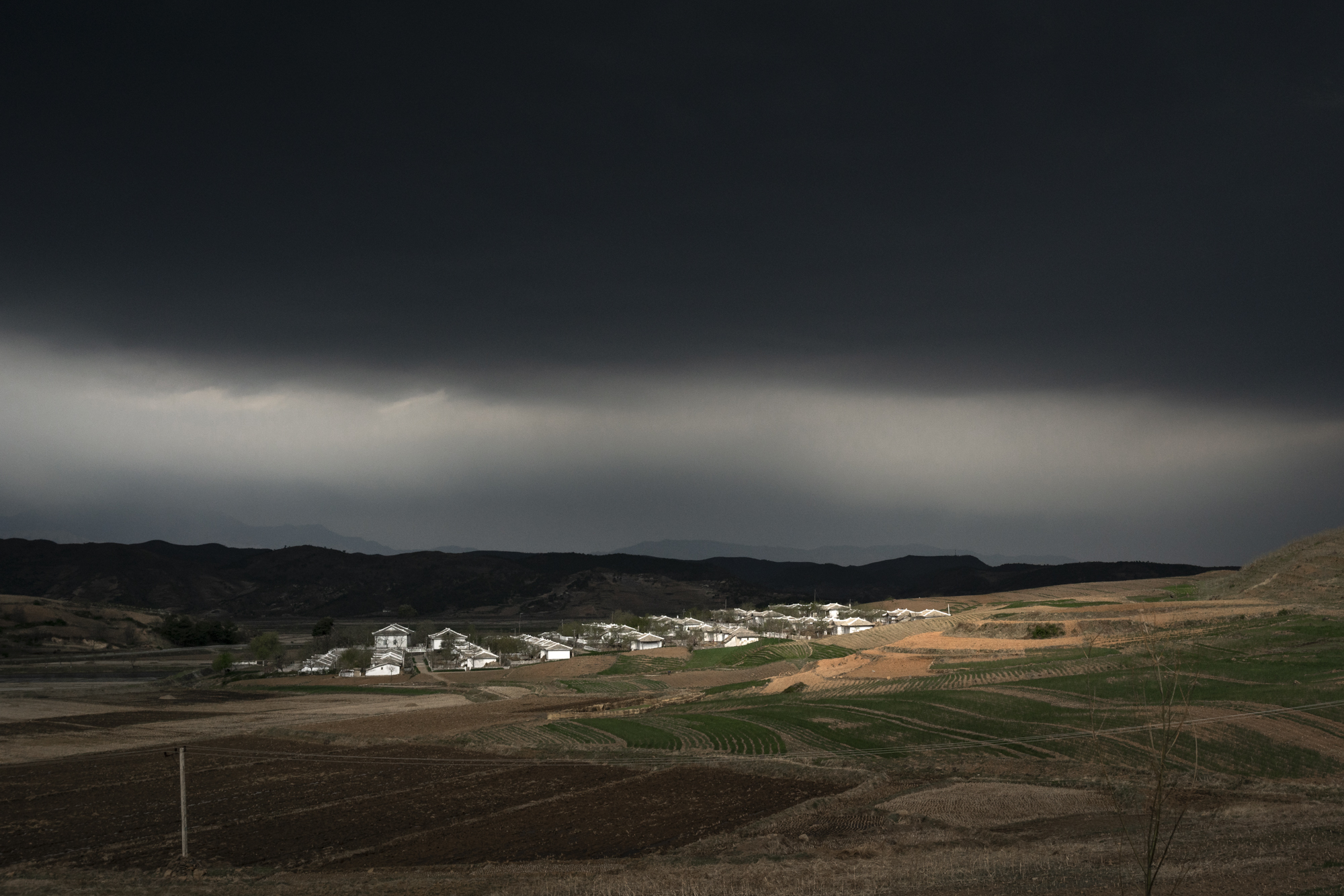  Under a gloomy cloud threatening rain, a beam of light shines on a small rural settlement in the Kangwŏn Province.  Forty percent of North Korea’s population lives in rural and agricultural communities. North Korean officials usually restrict foreig