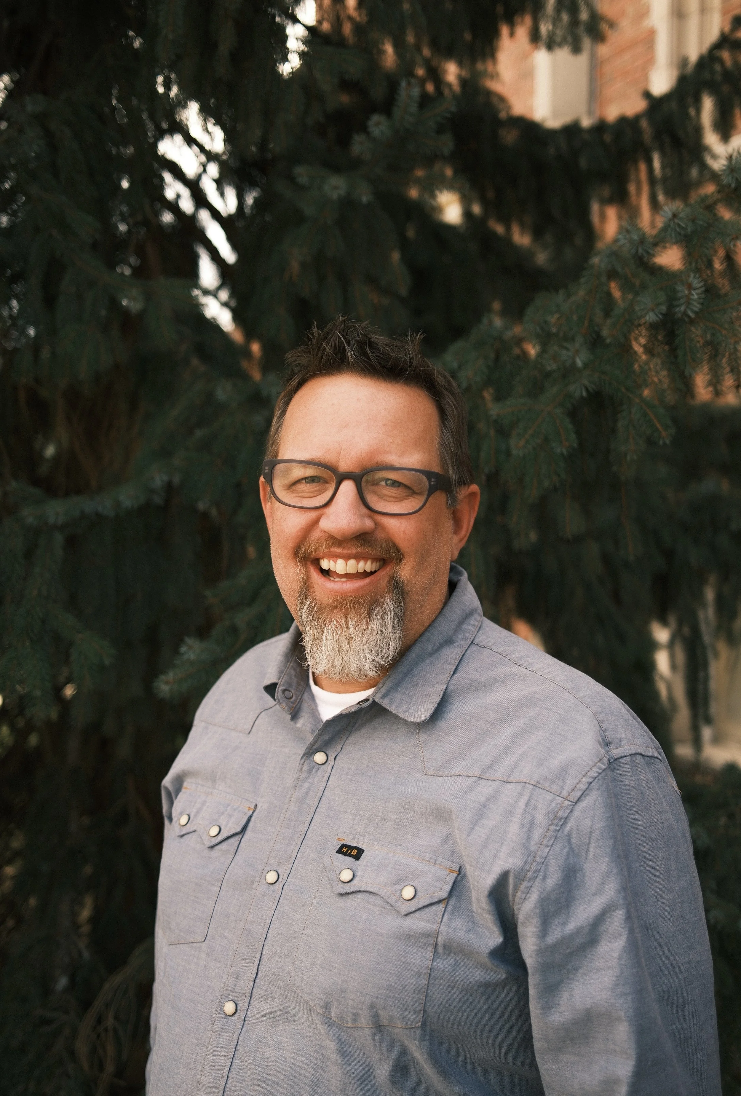 Portrait of a smiling man with glasses, a beard, and a mustache sitting in front of a large evergreen tree.