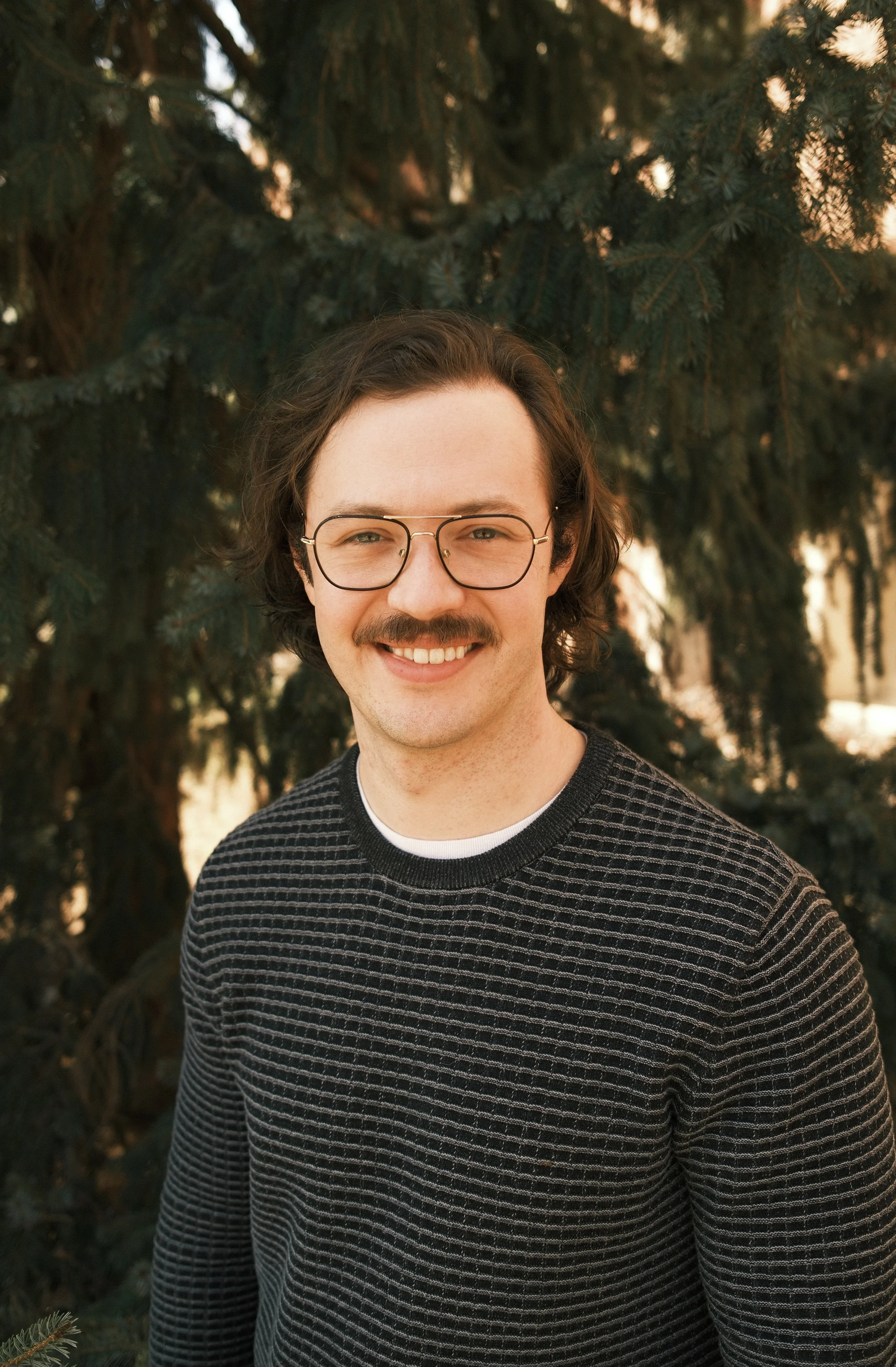 A young man with glasses and a mustache smiling outdoors in front of evergreen trees at sunset.
