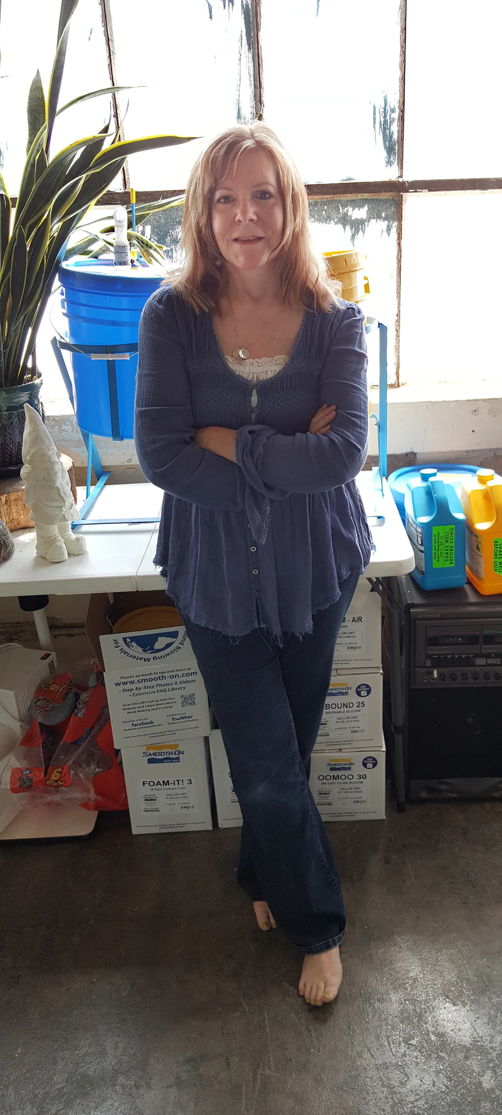A white woman with medium-length red hair in jeans & a blue top leans against a table in a cluttered studio.