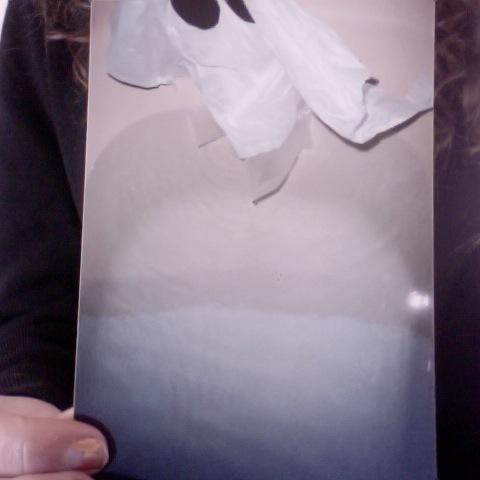 A snapshot showing a paper ghost emerging from a bathtub is held by a white woman obscured by the photo except for part of her hand, a black shirt, and curly brown hair.