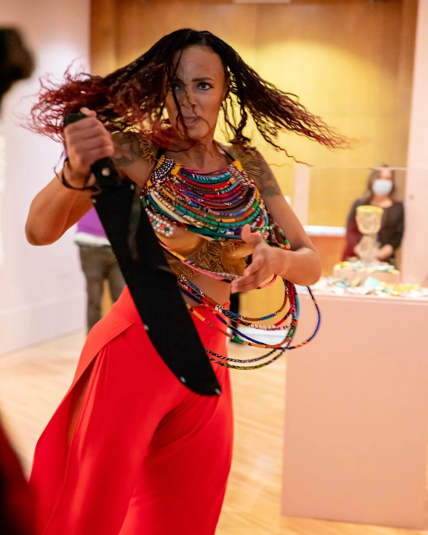 Caribbean woman wearing red pants, frozen mid-dance, wields a machete with focus. Her braided hair flies, and a vibrant, multicolored layered necklace adorns her, exuding command and vigor.