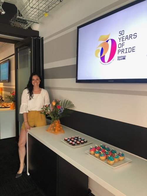 Image: Photo of an Edelman employee at their film screening in front of a TV screen that says "50 Years of Pride" and a table with food on it