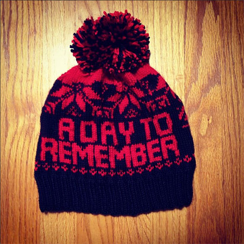 A red and black knit winter hat with a pom-pom on top, featuring a pattern of reindeer and snowflakes, and the words 'Always Remember' knitted into the design.