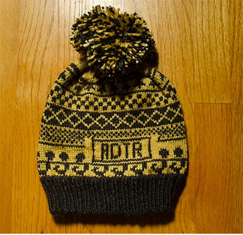 Yellow and black knit winter hat with a pom-pom on top, featuring the word 'NOTA' and decorative patterns, laid on a wooden surface.