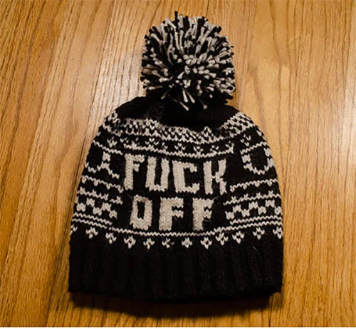 Black and white knit winter beanie hat with a pom-pom on top, featuring bold text saying 'FUCK OFF' and decorative snowflake and geometric patterns.