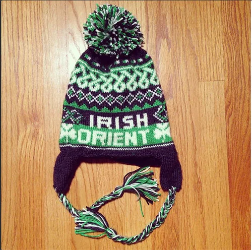 Knitted Irish-themed winter hat with a pom-pom on top, featuring green, purple, and black patterns, and tassels hanging from earflaps.