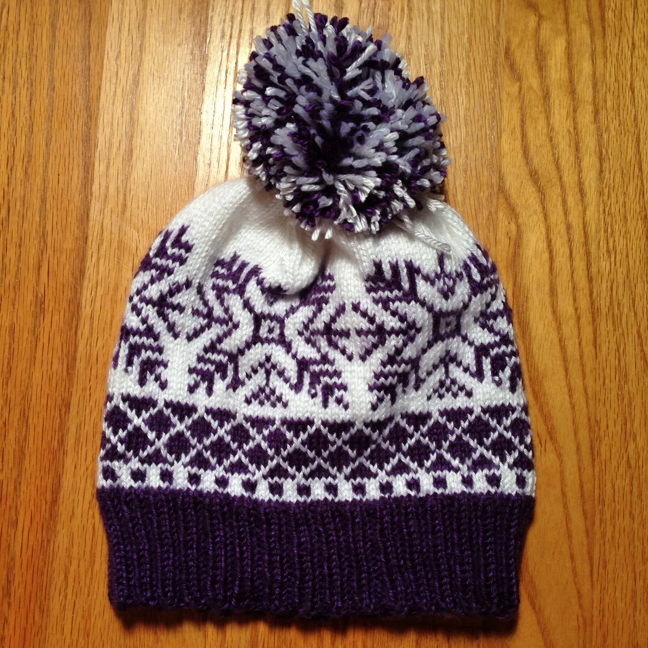 A knitted winter hat with a purple, black, and white snowflake pattern and a large pom-pom on top, placed on a wooden surface.