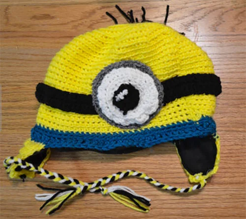 A crocheted Minion-themed hat with a yellow top, black eye band design, white eye with black pupil, and blue stripe, placed on a wooden surface.