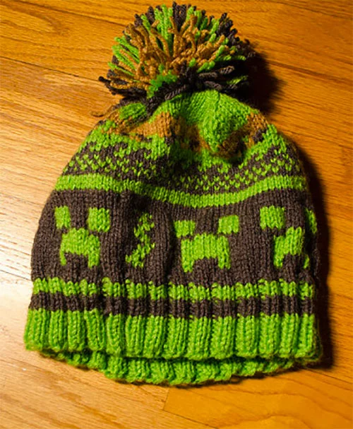 Green and black knitted beanie with alien spaceship and UFO designs, pom-pom on top, on a wooden surface.