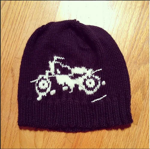 Black knit beanie hat with a white pixelated design of a motorcycle on it, placed on a wooden surface.