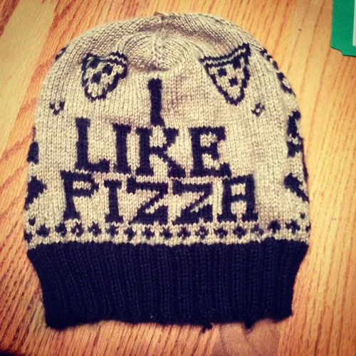 Gray knitted beanie hat with black lettering that says "I LIKE PIZZA" and various small heart and pizza slice patterns.
