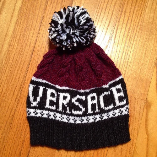 A knitted Versace winter hat with a pom-pom on top, colors of black, white, and red, lying on a wooden surface.