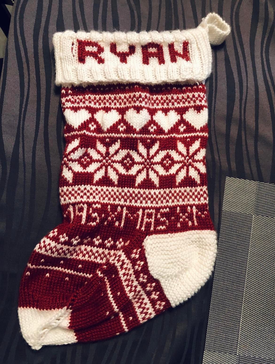 A Christmas stocking with red and white knit patterns, including snowflakes, hearts, and the name 'RYAN' at the top.