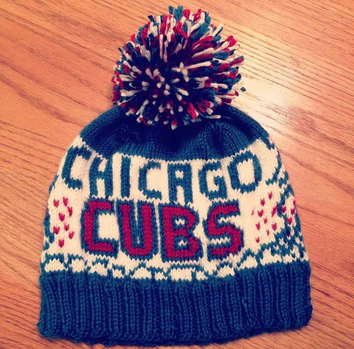 A knit winter hat with a pom-pom on top, featuring the words "CHICAGO CUBS" and decorative red and pink hearts.