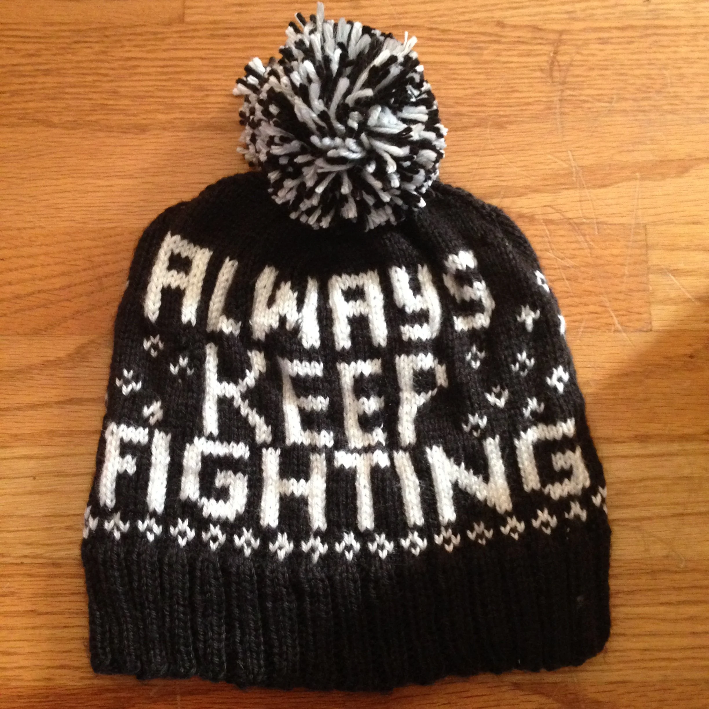 A black and white knitted winter hat with earflaps and a large pom-pom on top, placed on a wooden surface.
