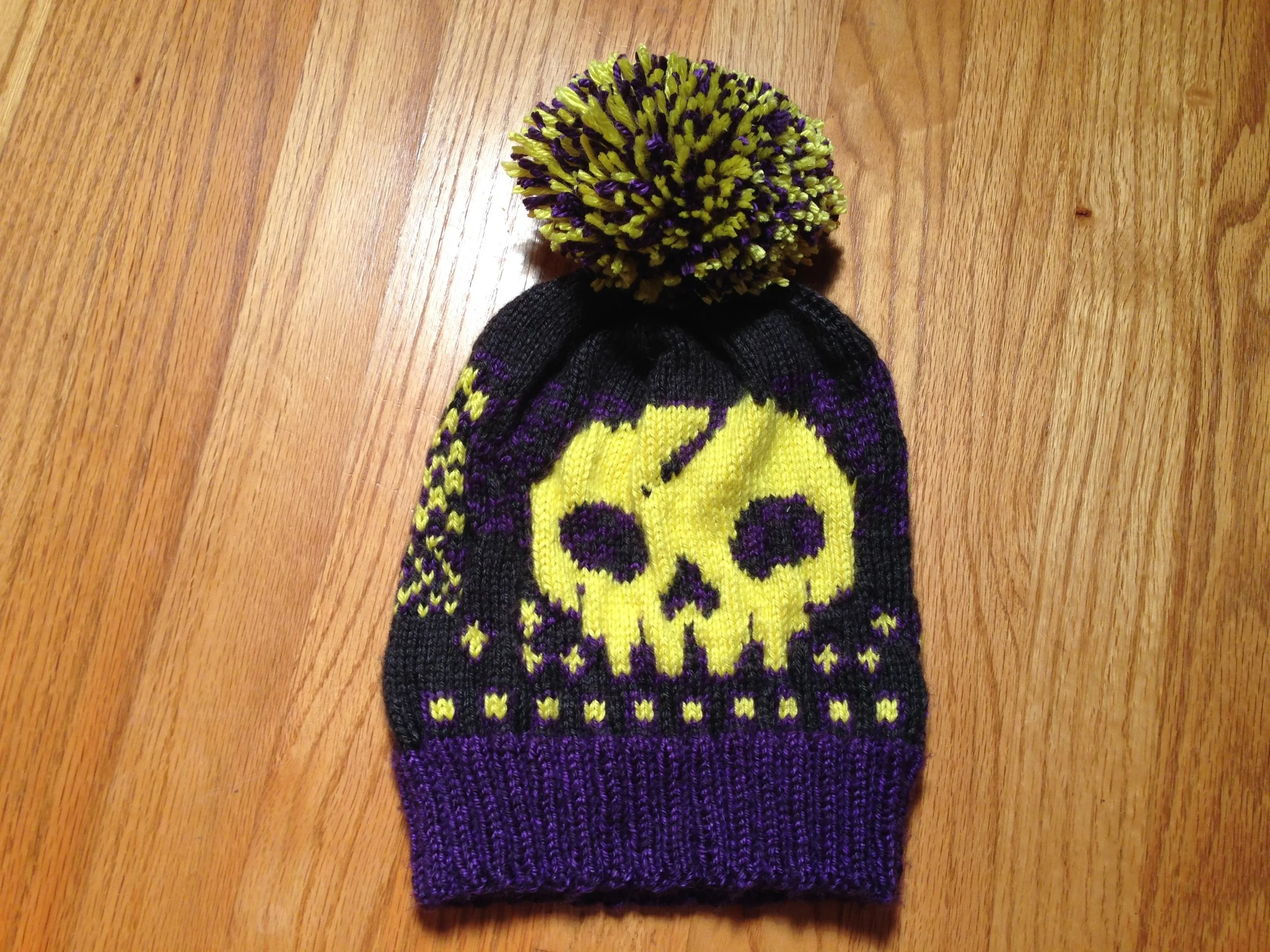 Close-up of a knitted winter hat with a yellow skull design, purple accents, and a large pom-pom on top, on a wooden surface.
