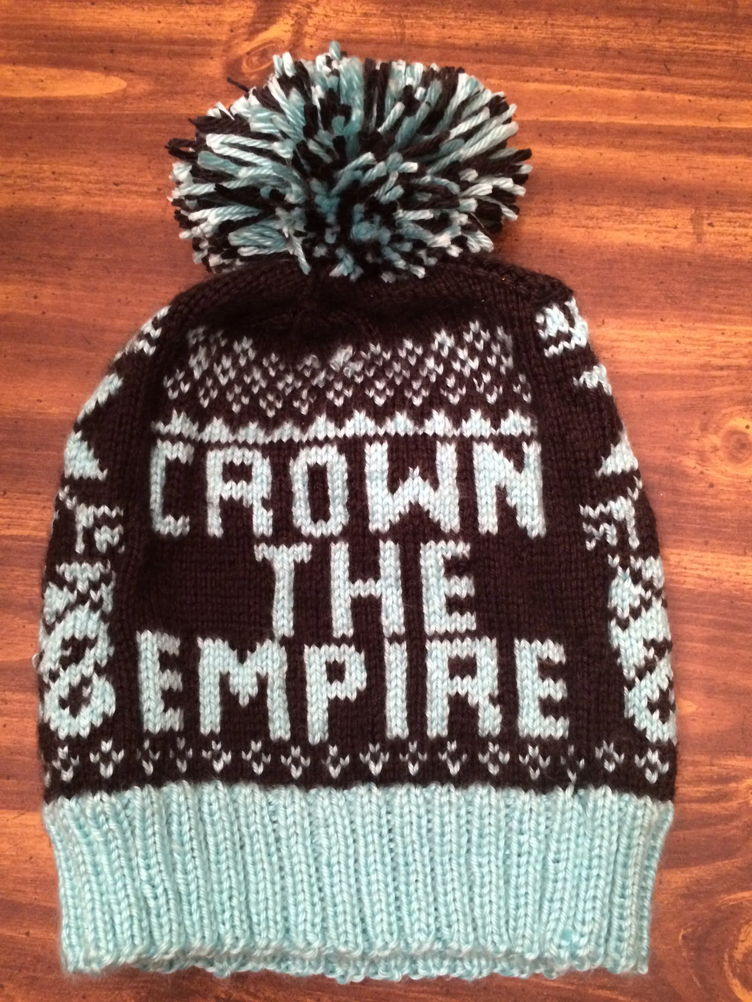 A knitted winter hat with black and light blue colors, featuring a pom-pom on top, lying on a wooden surface. The hat has a pattern with the words 'CROWN' and 'KING' in white letters.