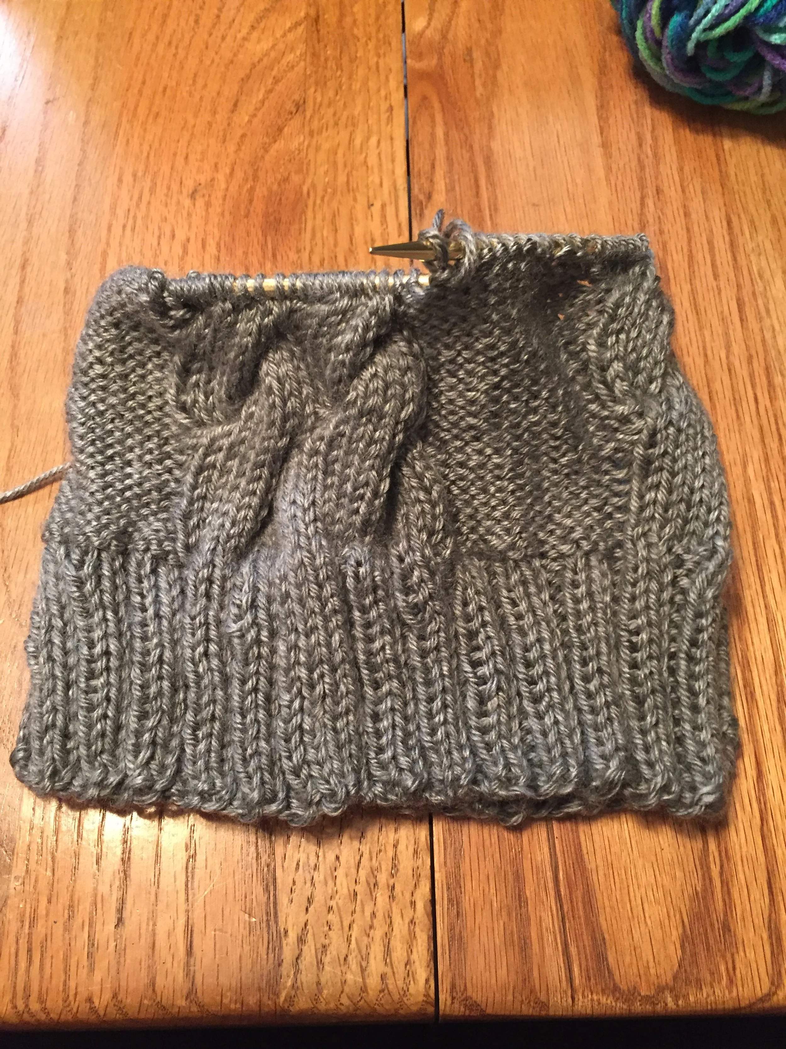 A piece of gray knitted fabric in progress on a knitting needle, resting on a wooden surface.