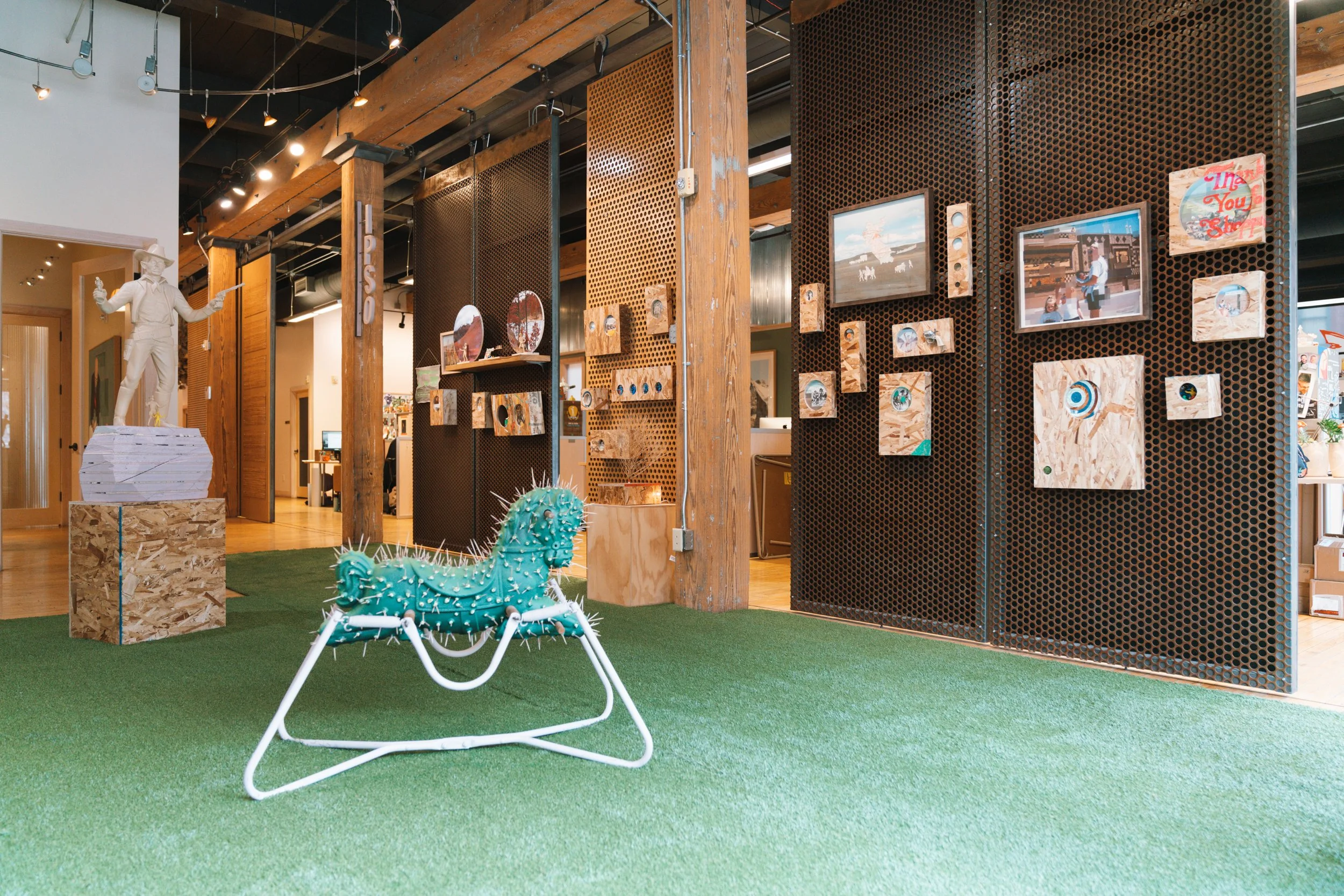 Indoor art exhibit with a white statue of a person in a cowboy hat pointing to the sides, a cactus-shaped sculpture, and a wall with framed art and photographs. The floor is green artificial turf and the space features exposed wood beams and black pe