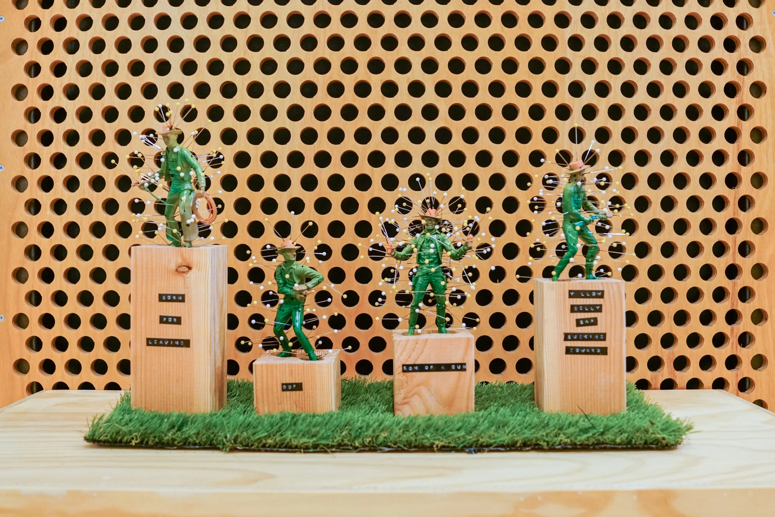 Display of four miniature green soldiers with pea shooters, each on a wooden pedestal with labeled captions, set on a grass patch against a wooden panel background with circular holes.