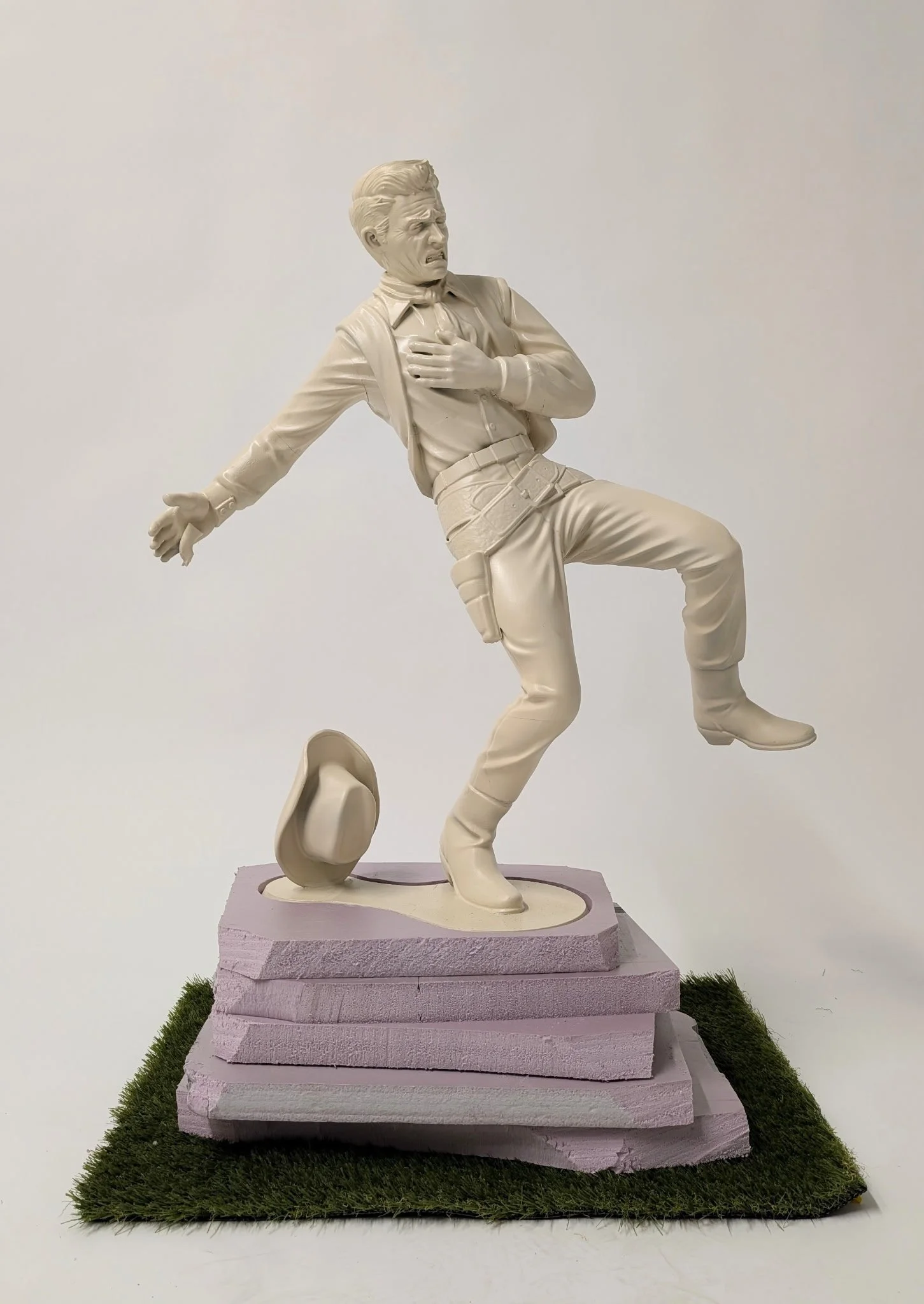 White sculpture of a man in a dynamic pose, holding his chest and one arm extended behind, standing on pinkish steps with artificial grass around base.