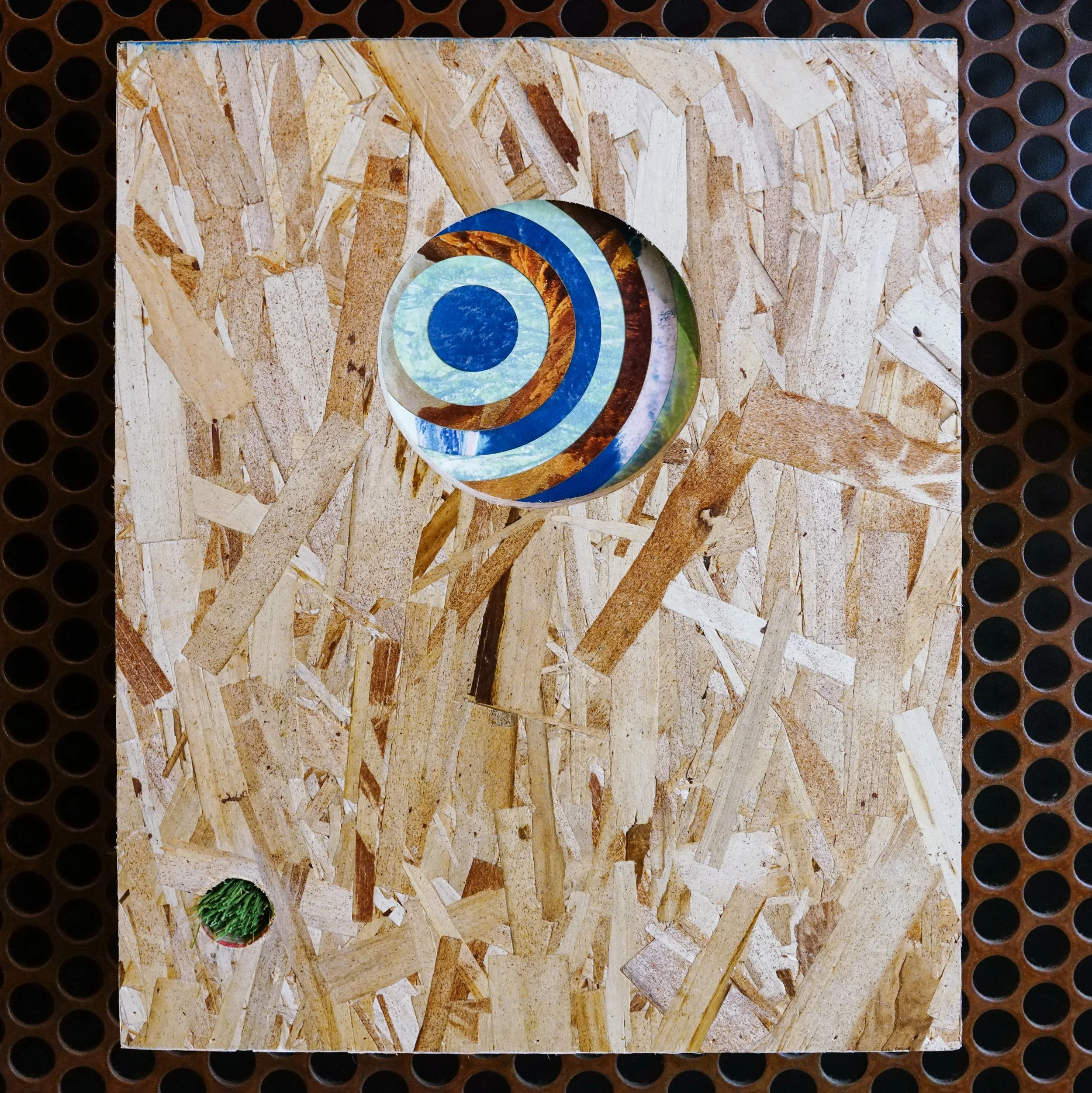 Close-up of a corkboard surface with a wooden spiral puzzle and a small cluster of artificial green grass in the bottom left corner.