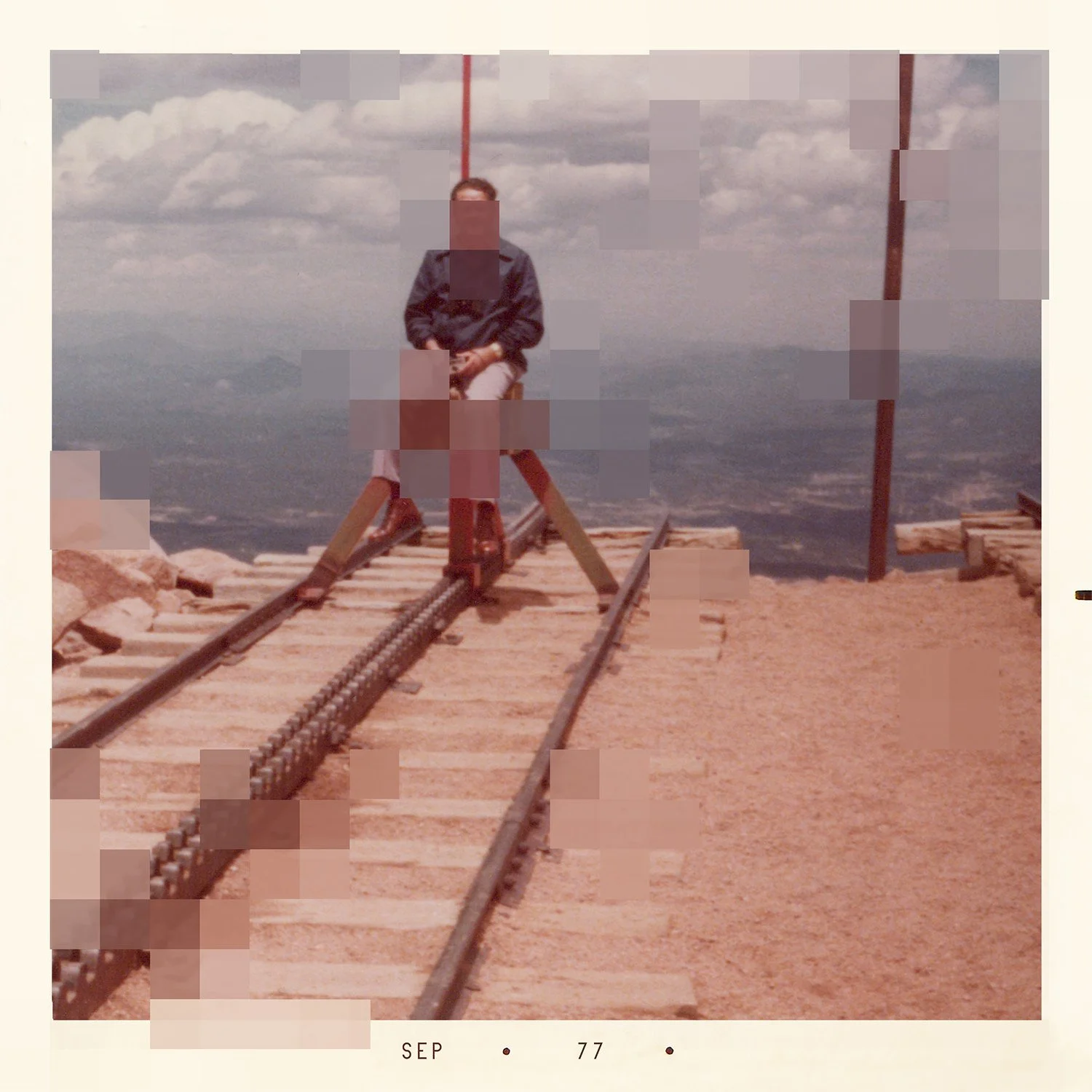 A person sitting on a wooden platform on a mountain top with train tracks in front, mountains in the background, and a cloudy sky overhead.