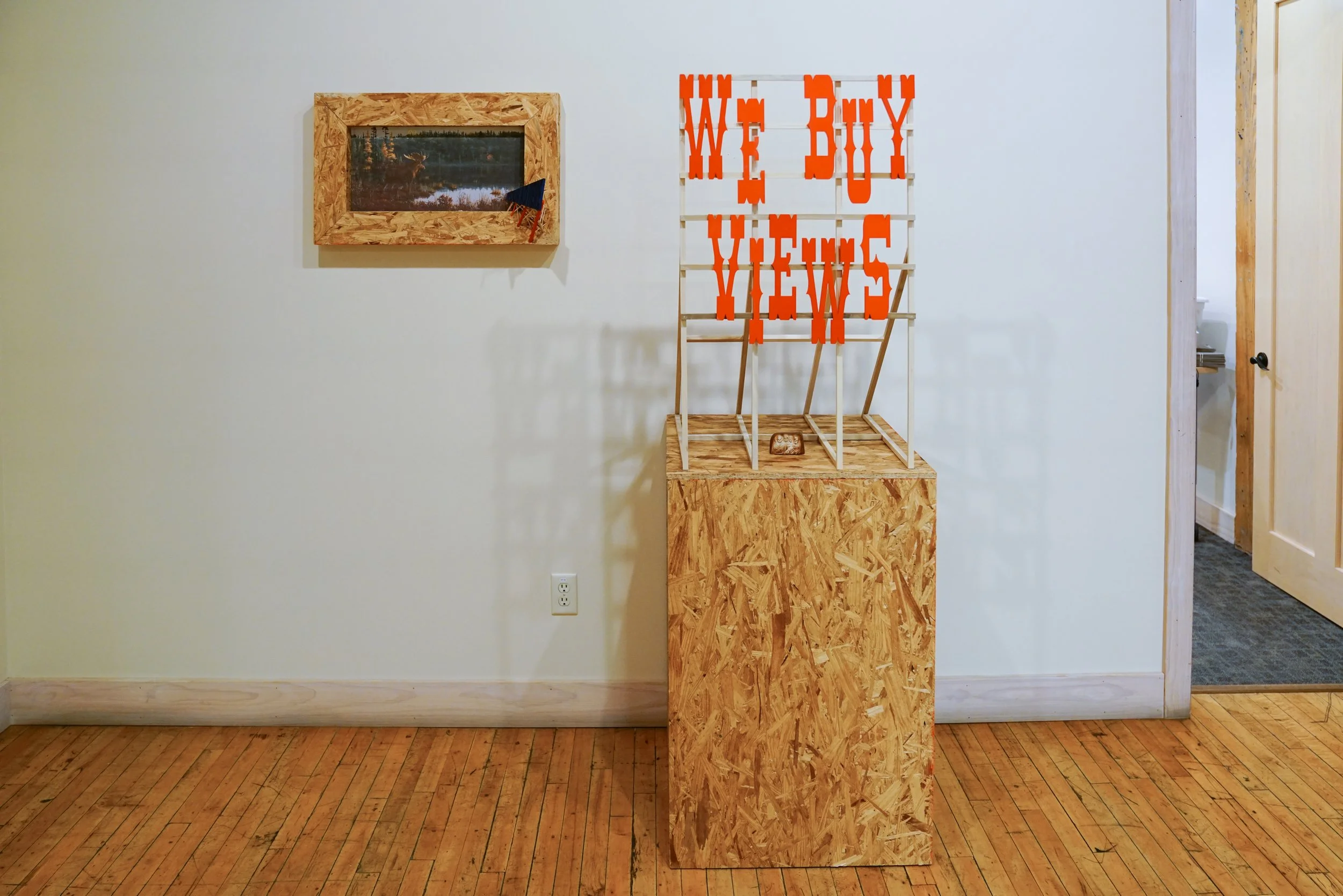 An indoor art exhibit displays a wooden structure with orange text saying "WE BUY MY VIEWS" on a white background frame.