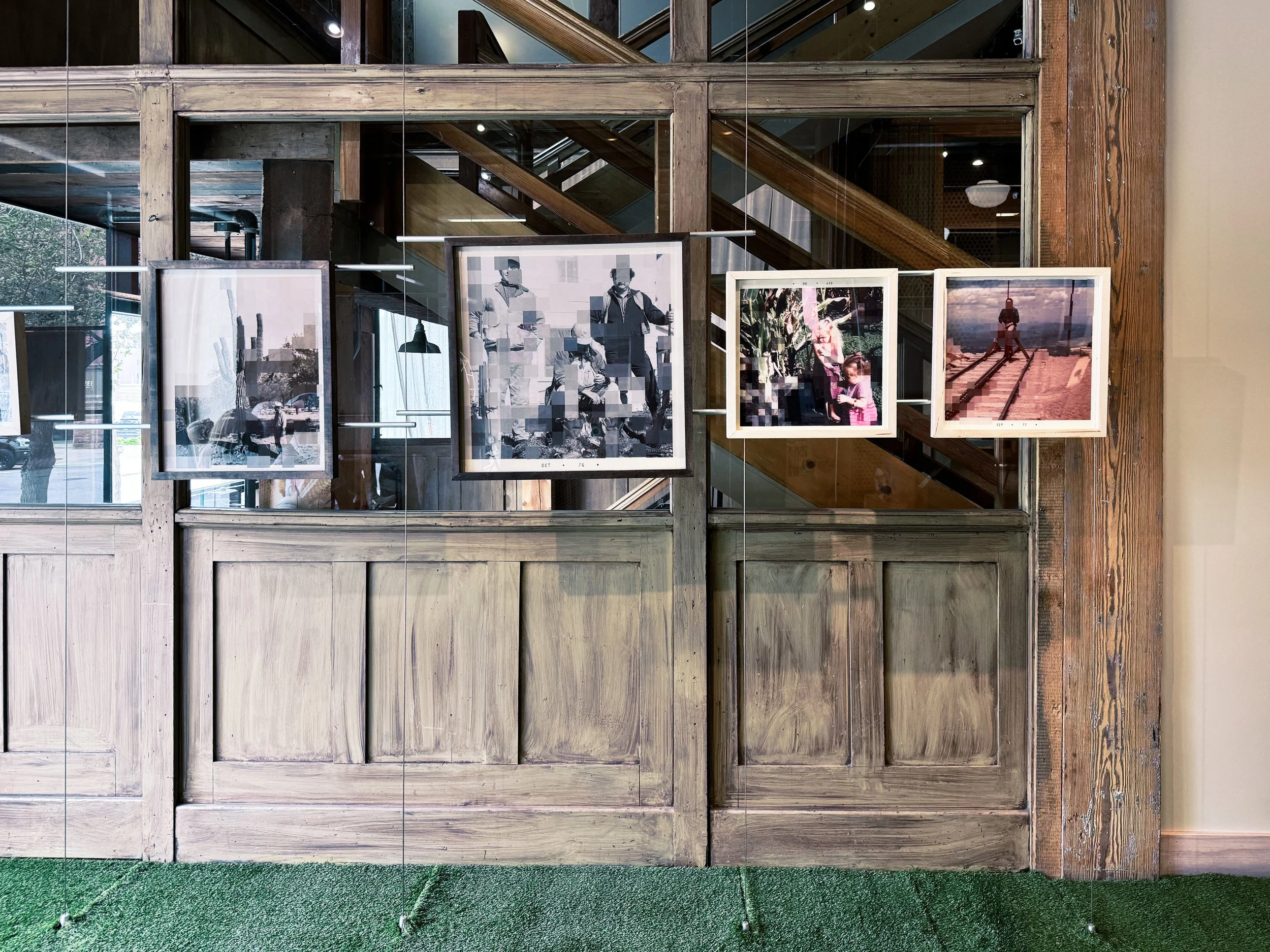Wooden indoor gallery with framed black-and-white photographs, green carpet, and staircase reflected in glass window.