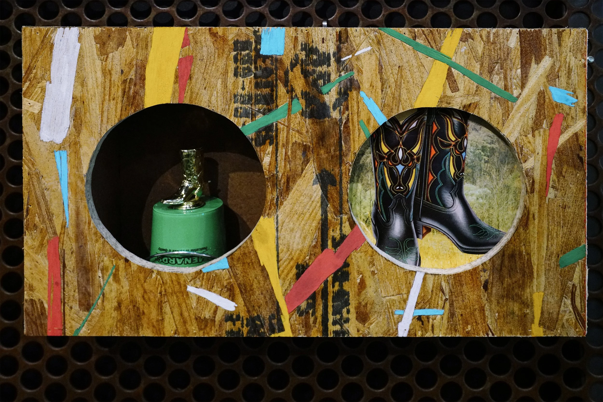 A wooden board with colorful painted lines and cutout holes, one of which contains a green plastic bottle with a silver cap and some greenery inside, and the other displaying a black and yellow patterned cowboy boot.