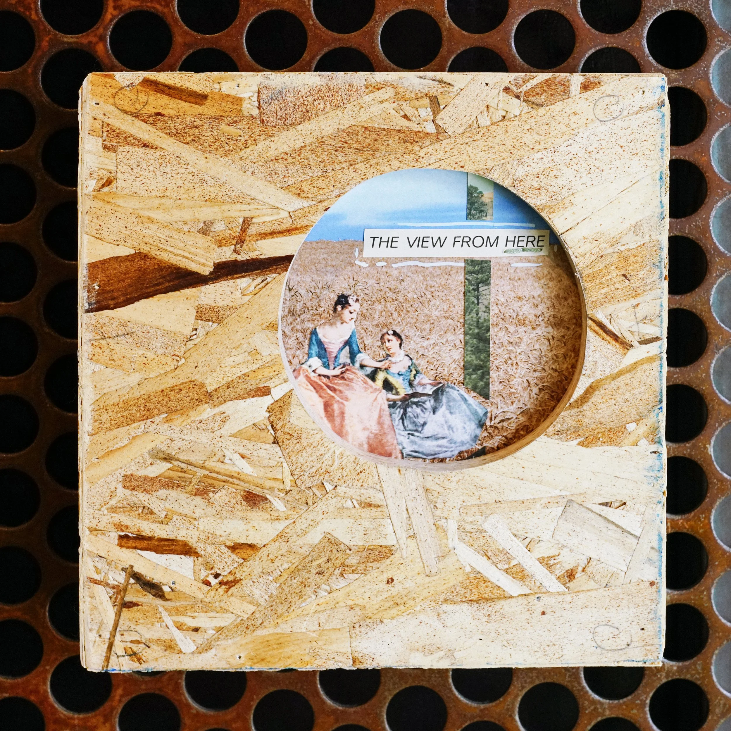 Close-up of a wooden plaque or sign with a photo collage and the partial text 'THE VIEW FROM HERE' on it, placed on a surface with circular holes or spots.