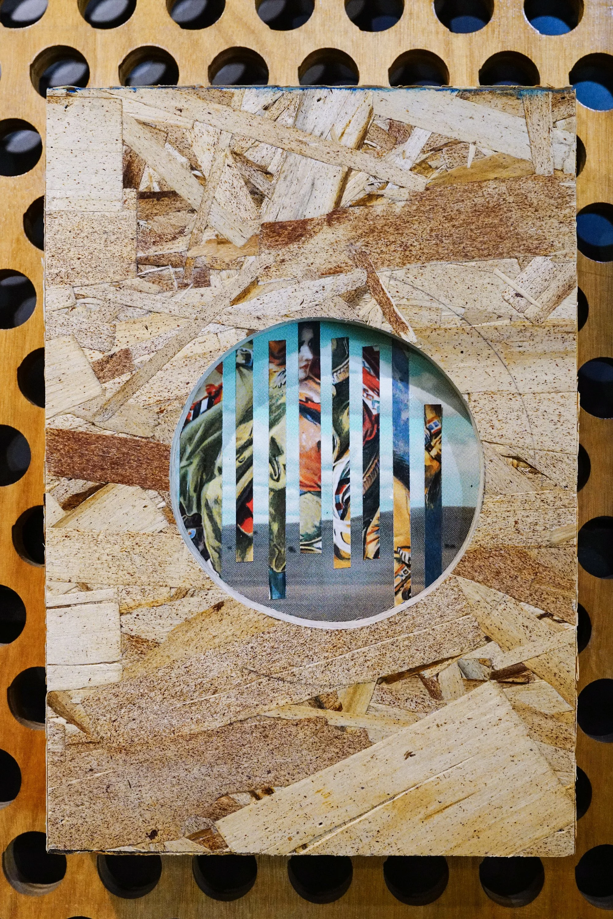 A close-up view of a vintage-style wooden speaker grille with a round cutout, showing a colorful image of motocross racers through the grille slats. The background features a wooden surface with circular holes.