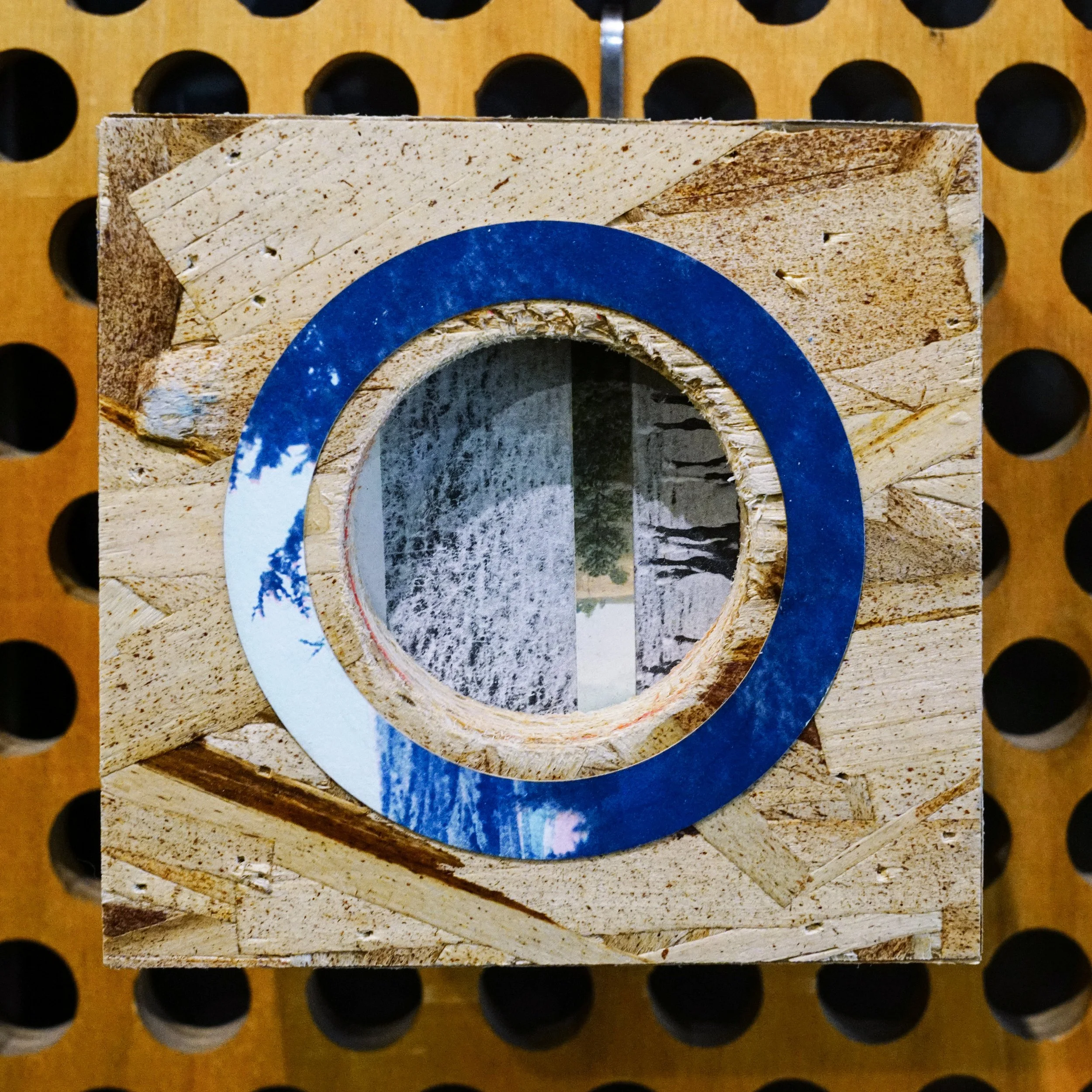 Close-up of a cardboard box with a circular hole and blue paint on the edge, placed on a perforated wooden surface.