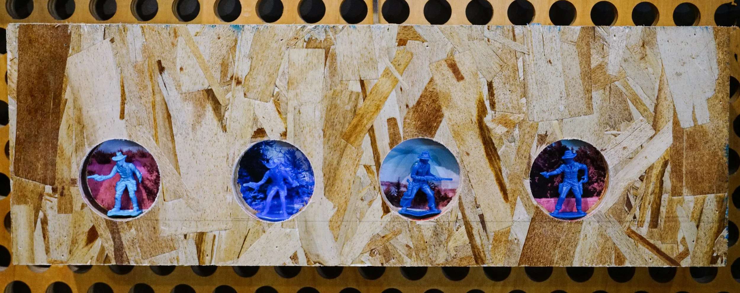 A wooden board with four circular cutouts, each containing a miniature soldier figurine painted blue and posed in various military stances.