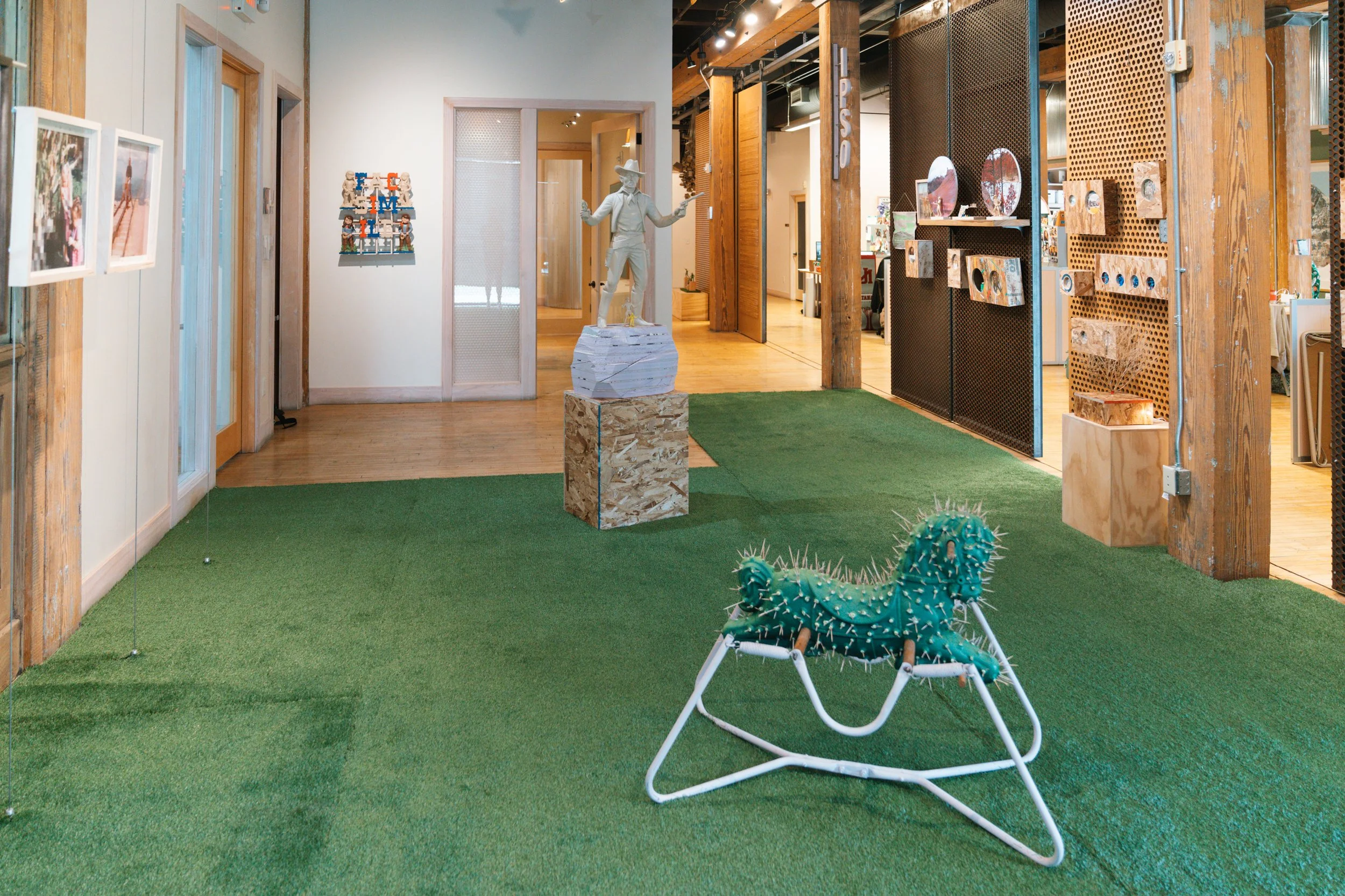 An indoor space featuring green artificial turf flooring, a cactus-shaped lounge chair with spikes, a white figurine of a cowboy or rancher with a hat and boots, wooden and metal decor elements, and artwork on the walls.