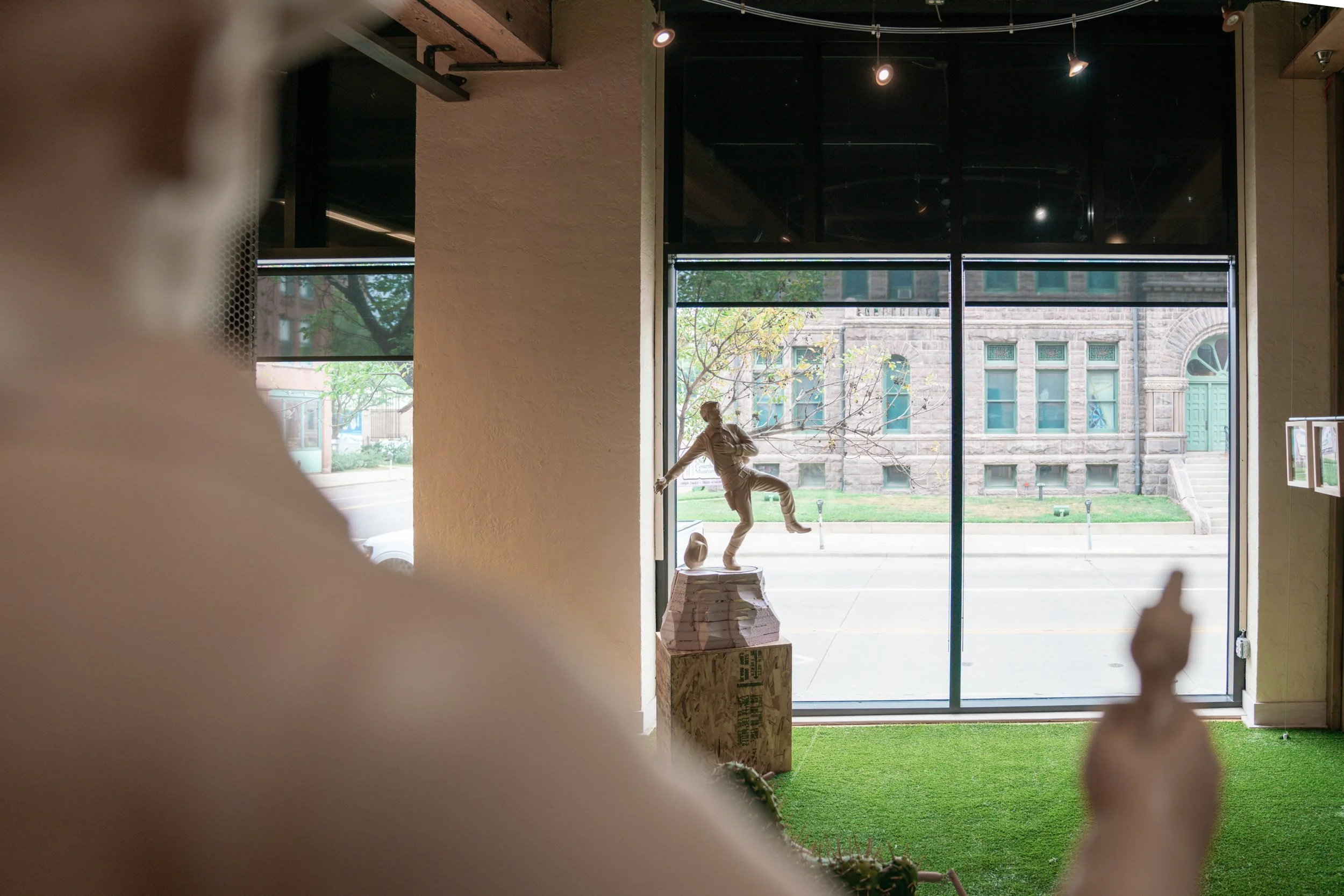 View from inside a building looking out through large windows onto a city street with trees and a historic stone building. Inside, there is a sculpture of a person, a cardboard box, and blurred fingers or hands in the foreground.