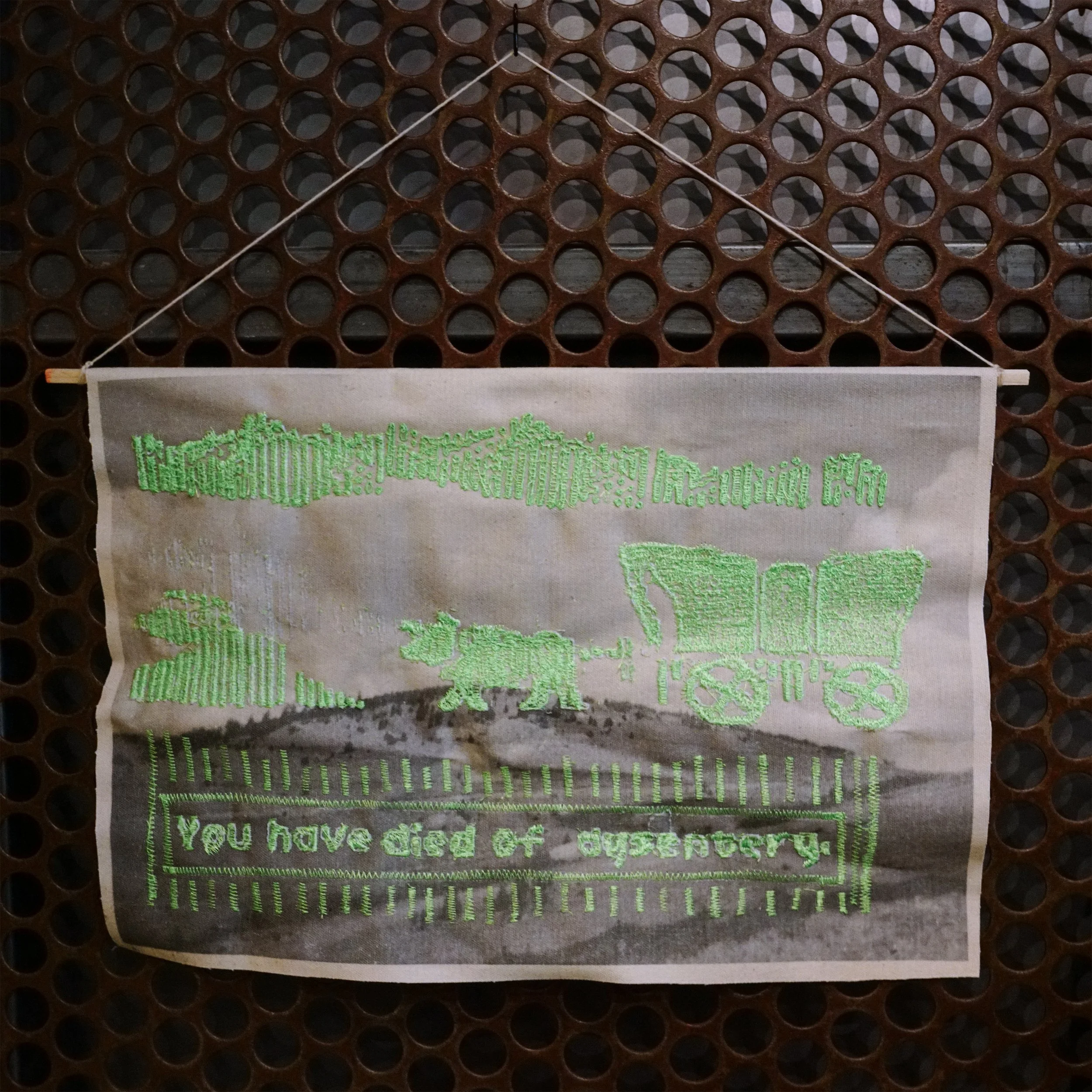 Close-up of a decorative wall hanging with embroidered text in green, against a brown, honeycomb-patterned background. Part of the text reads "You have died of...".