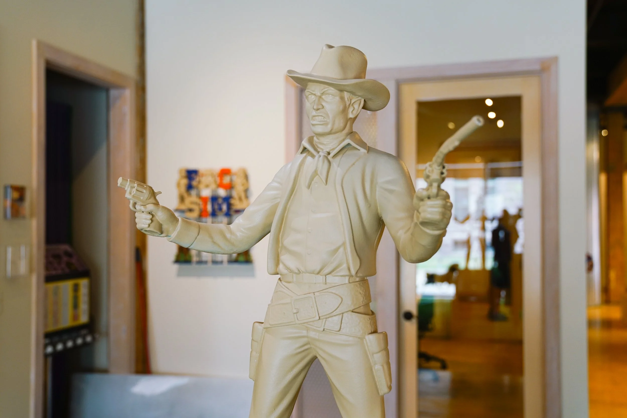 A white sculpture of a cowboy holding a gun and a lasso, wearing a hat and bandana, situated indoors with rooms and walls visible in the background.