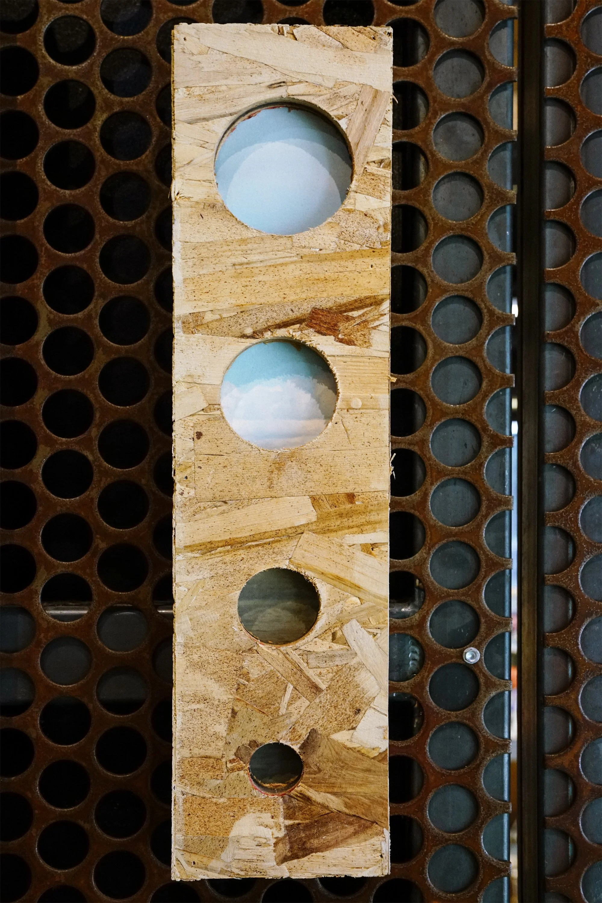 A piece of oriented strand board with four circular holes placed vertically, set against a background of a brown perforated metal sheet.