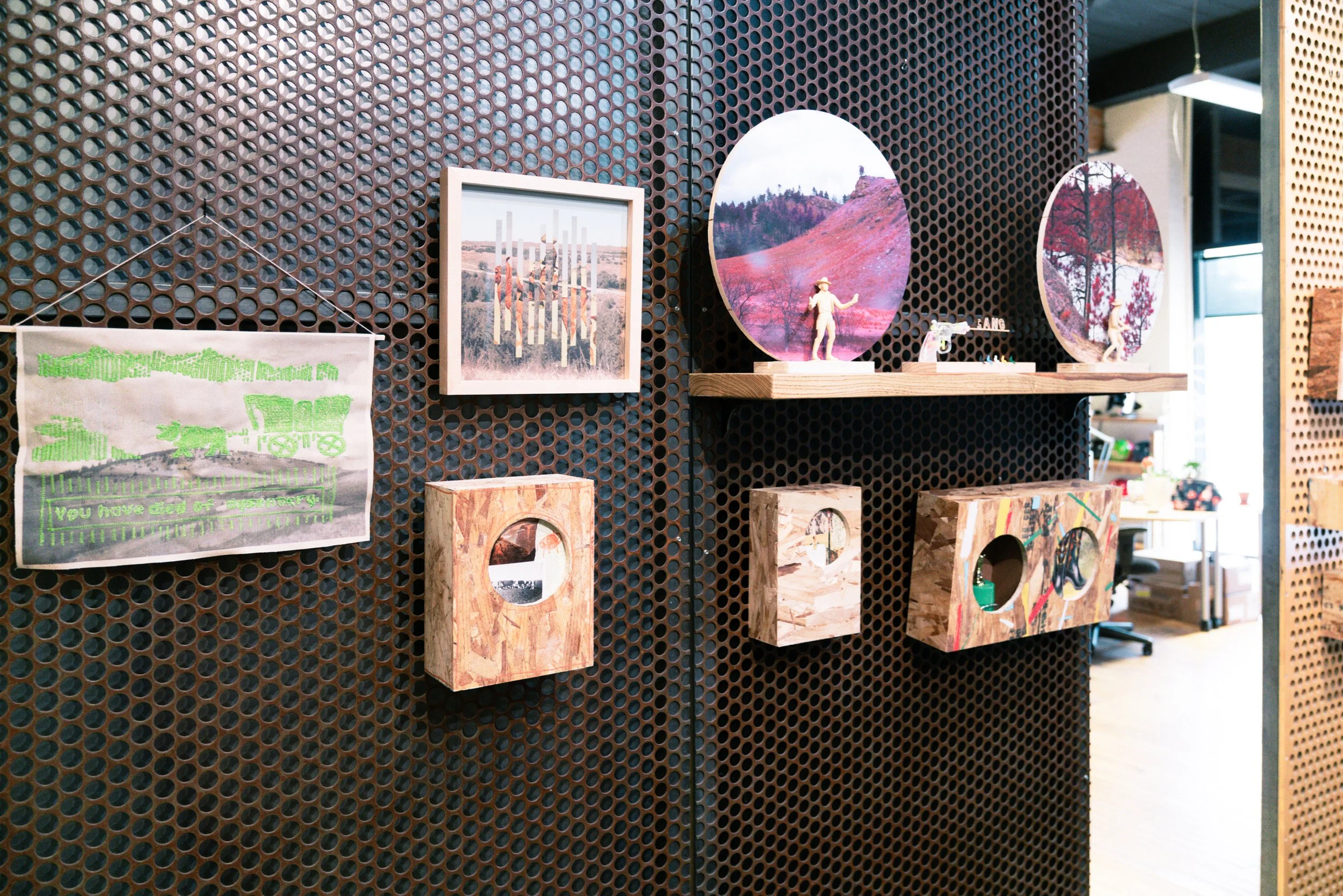 Display wall with various framed artworks, including landscape photos, small sculptures, and wooden blocks on a metal grid background.