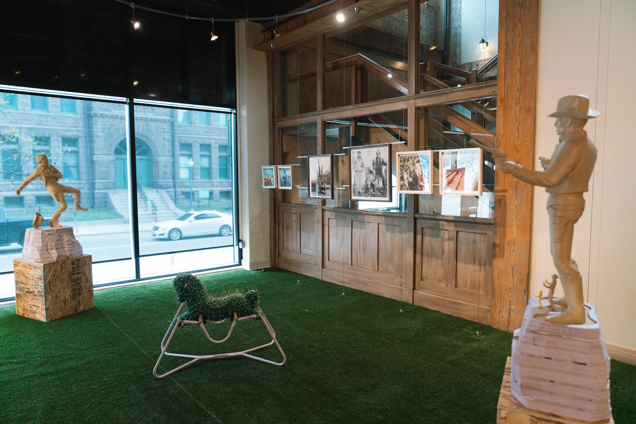 Art gallery with framed photographs on a wall, sculptures including a running figure, a cactus, and a wire chair, with large windows showing a city street outside.