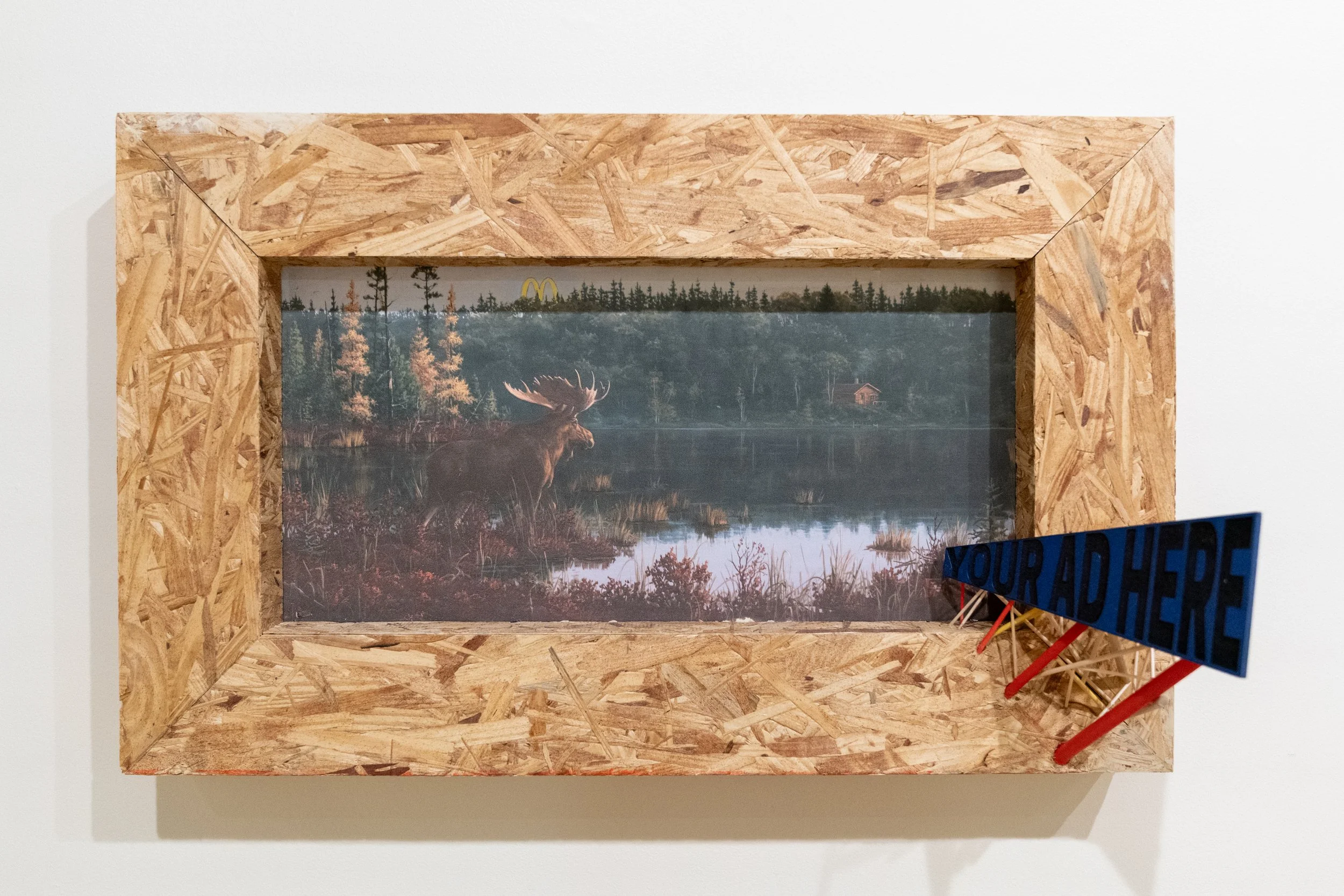 Framed landscape artwork of a moose in a forested lake scene with a distant house and an overcast sky, set within a wooden frame on a white wall, a blue sign with red legs reading 'Your Advice?' is in the foreground.