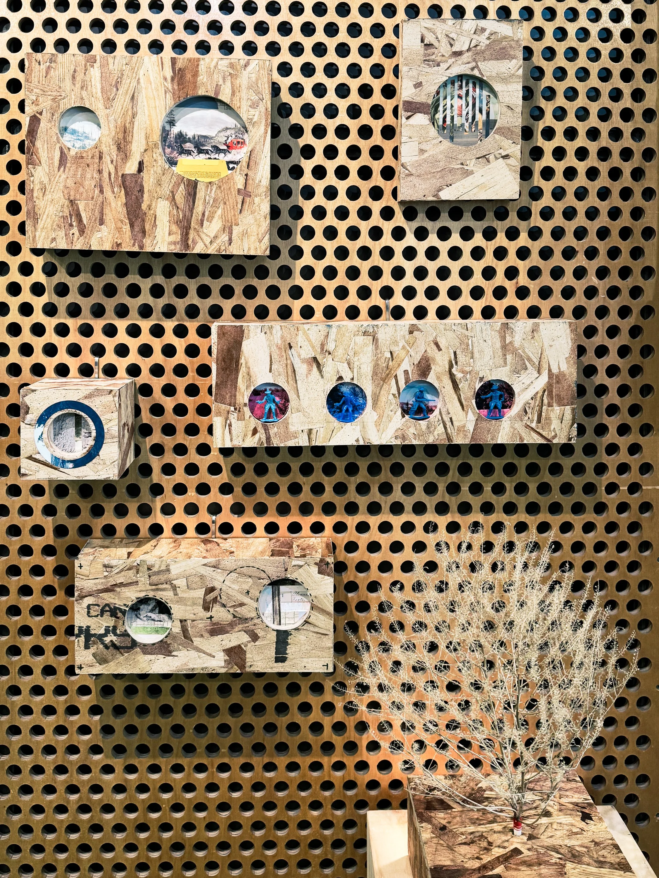 Display of various wooden art pieces with circular cutout windows showing different scenes and textures, arranged on a board with a perforated background and some dried plant decoration.